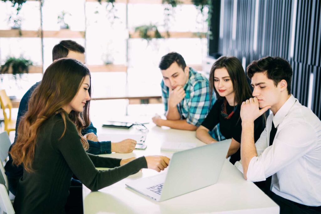 Five co-workers sitting at a table looking at a laptop during a presentation