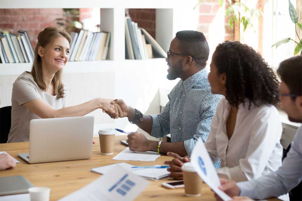 Four business people sitting at a table talking, two of them shaking hands and smiling