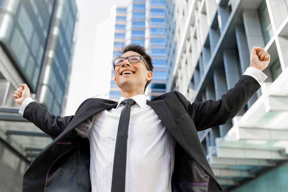 Happy businessman outside highrises in a city celebrating with his fists raised in triump