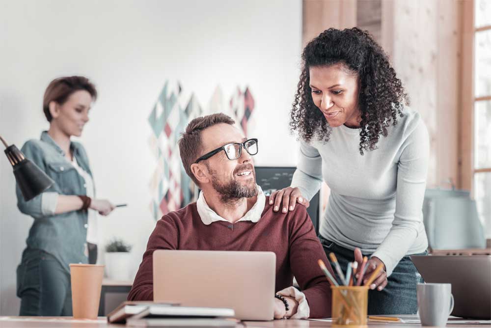 A coworker seated at a desk with a laptop with a co-worker standing next to him with her hand on his shoulder